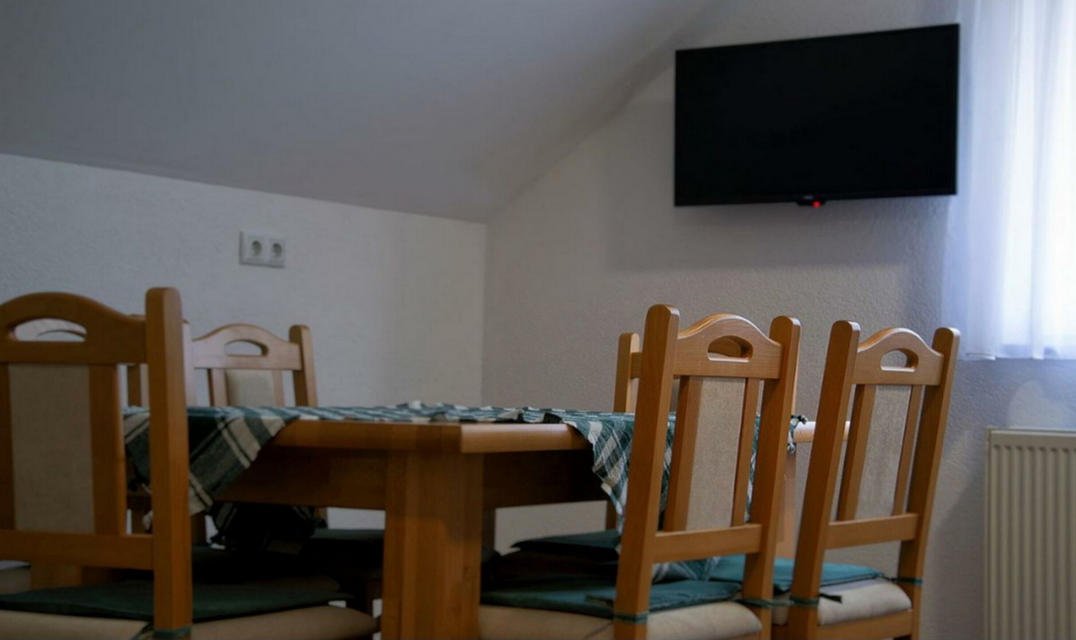 Un área de comedor con una mesa y cuatro sillas de madera, un televisor de pantalla plana en la pared y un ventanal con cortinas.