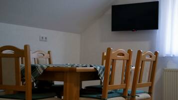 Un área de comedor con una mesa y cuatro sillas de madera, un televisor de pantalla plana en la pared y un ventanal con cortinas.