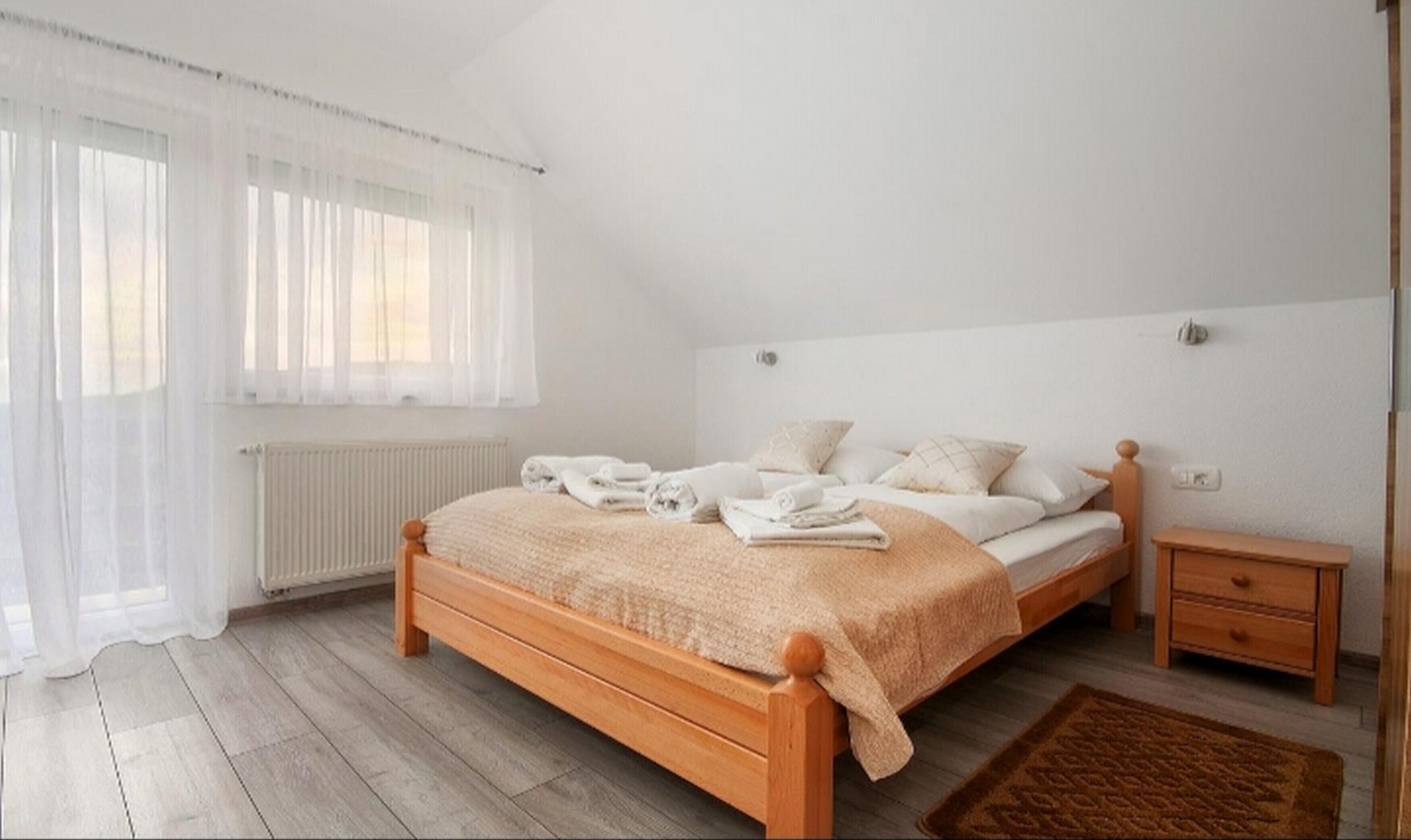 A bedroom with a wooden bed, white bedding, a bedside table, and a window with sheer curtains.
