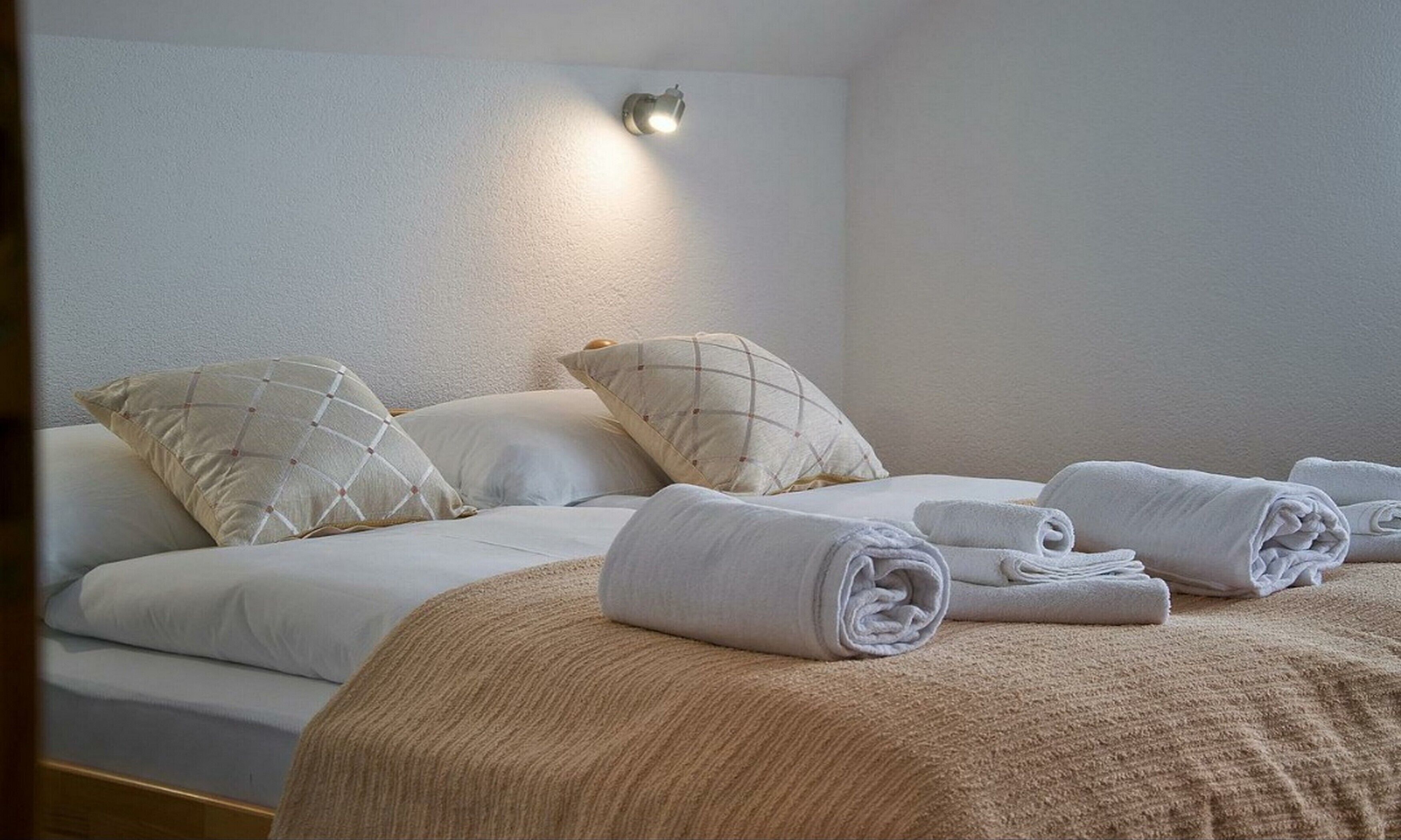 Una cama bien tendida con un edredón color beige, dos almohadas con estampado de cuadros y toallas enrolladas sobre la cama.
