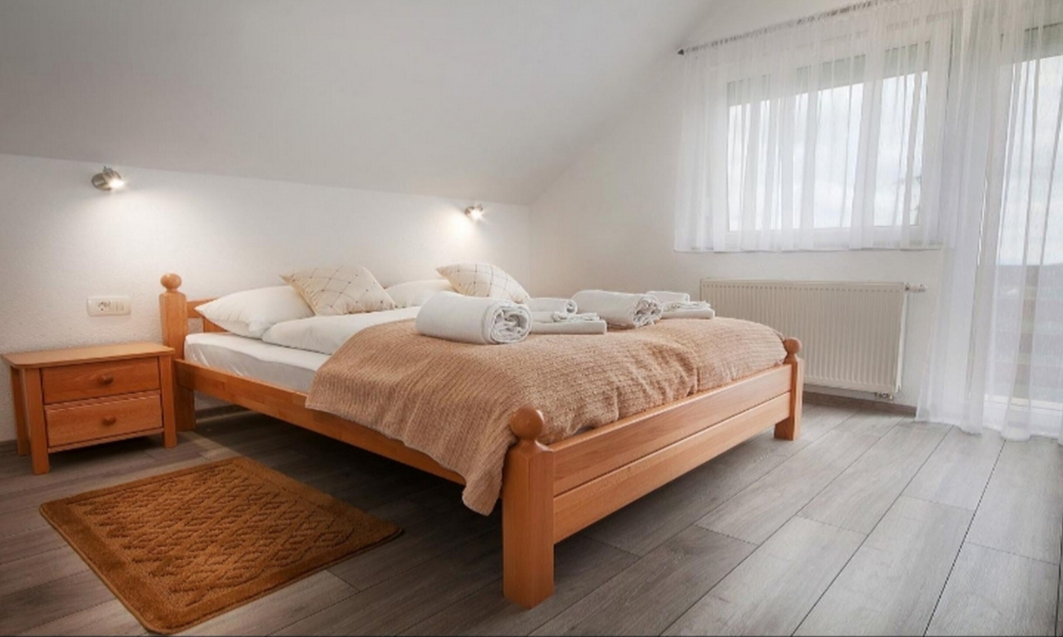 Un dormitorio con cama de madera, ropa de cama blanca, una mesita de noche, una alfombra marrón y una ventana con cortinas de tul.