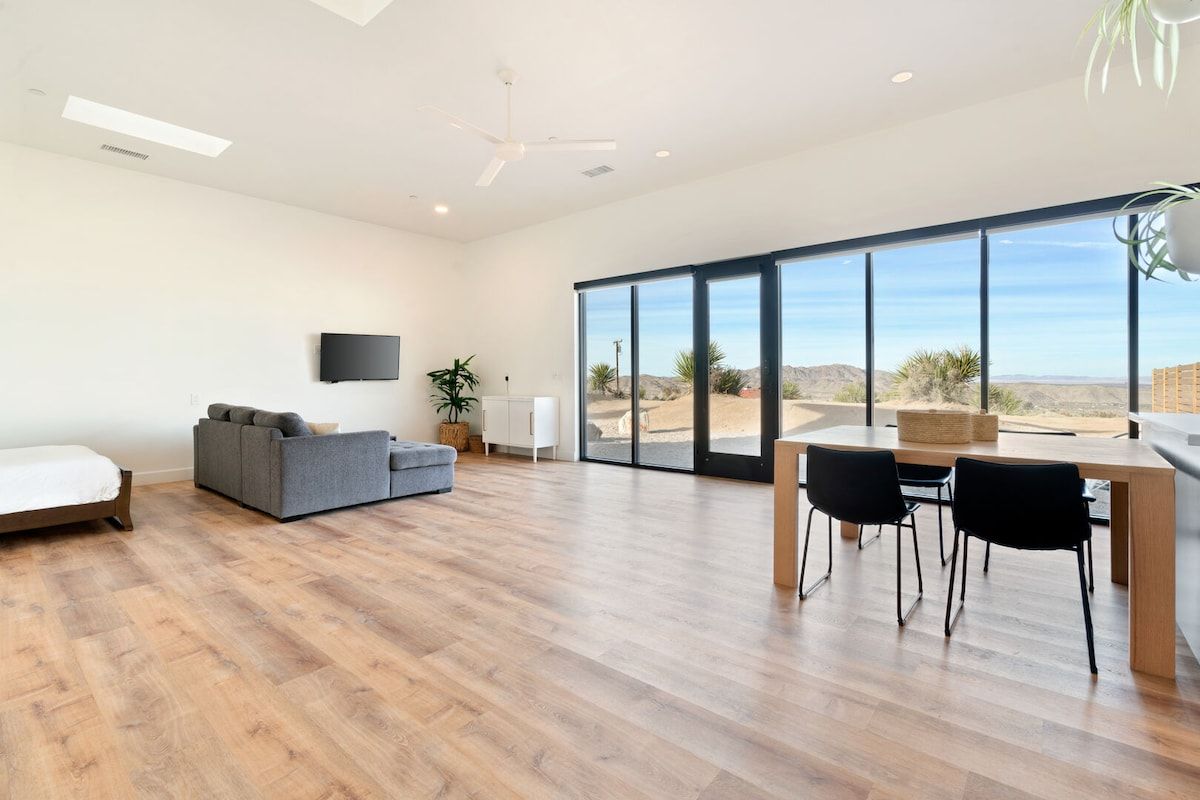 A spacious room with a wooden floor, a gray sofa, a dining table with chairs, and a large window offering a view of the desert.