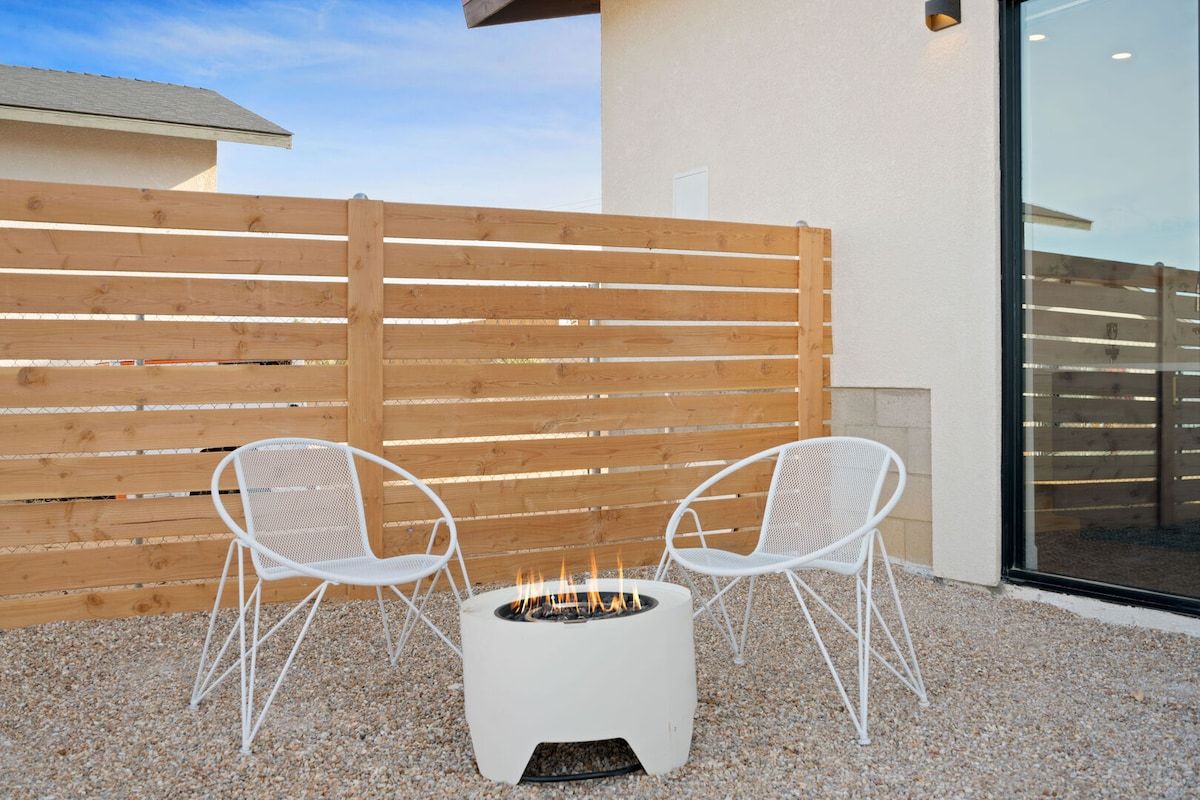 A fire pit with two white chairs in a gravel area, a wooden fence, and a building with a glass door.