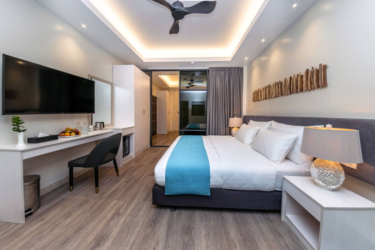 Een moderne hotelkamer met een flatscreen-tv, een bureau met stoel, een bed met blauwe deken, een nachtkastje met lamp en een plafondventilator.