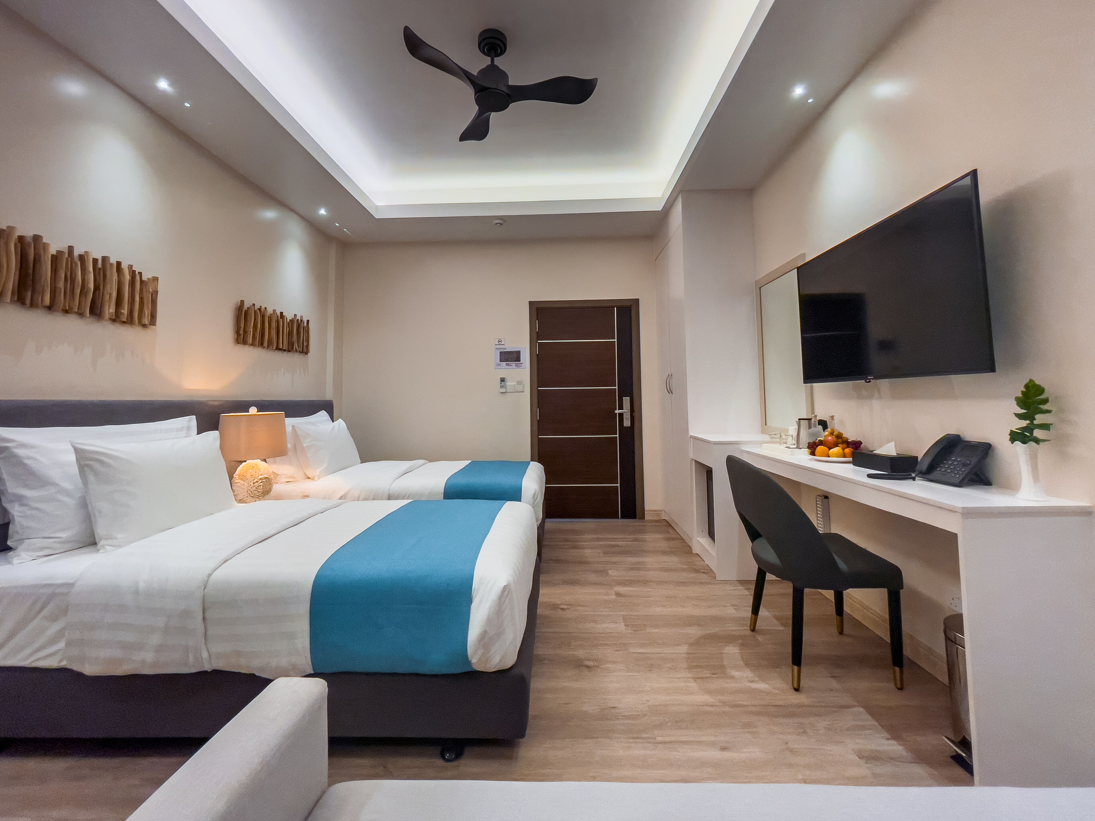 Une chambre d’hôtel avec deux lits, un bureau, une chaise, un téléviseur à écran plat et un ventilateur de plafond.