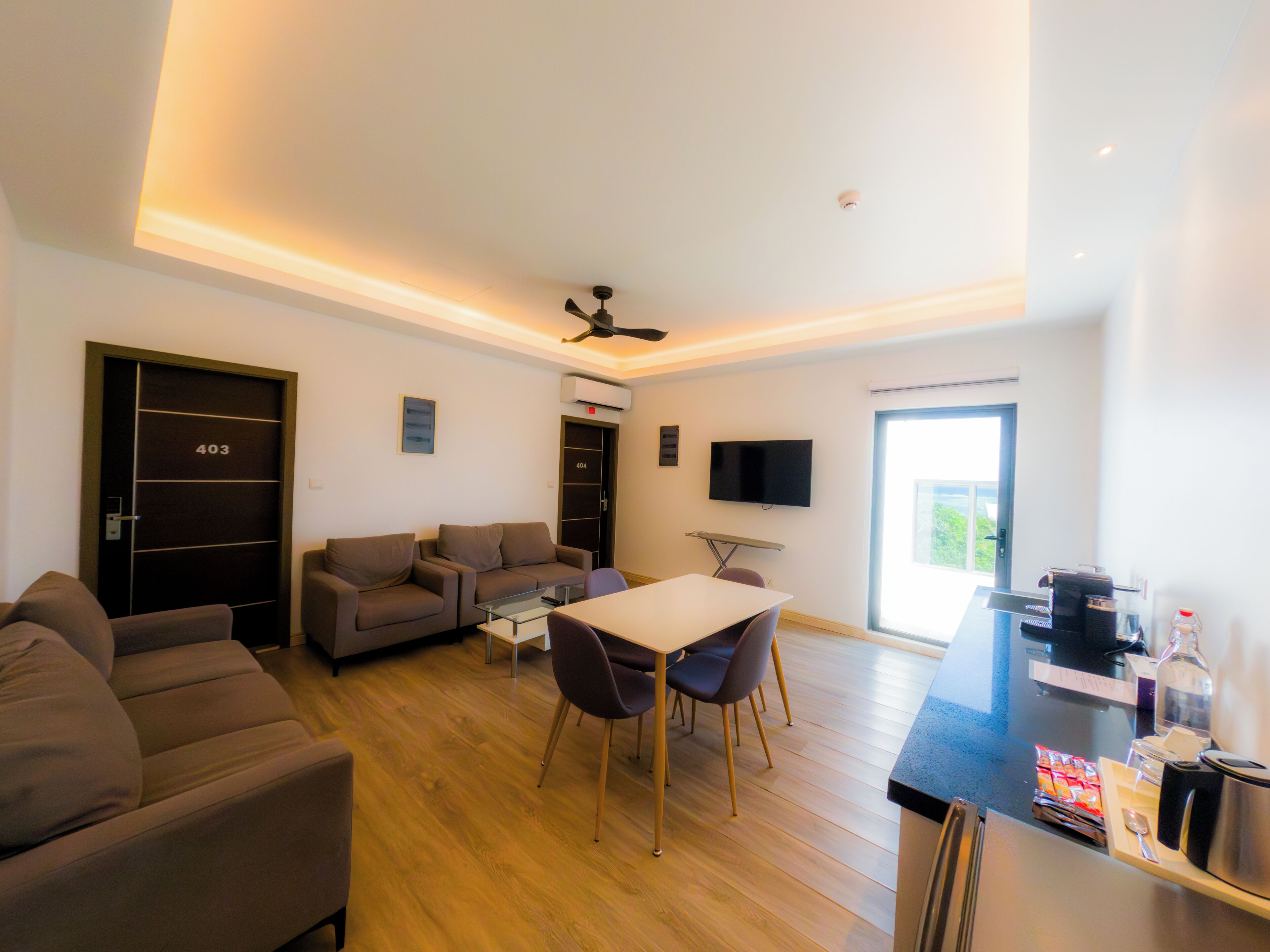 A modern living room with a sofa, a dining table with chairs, a flat-screen TV mounted on the wall, and a ceiling fan.