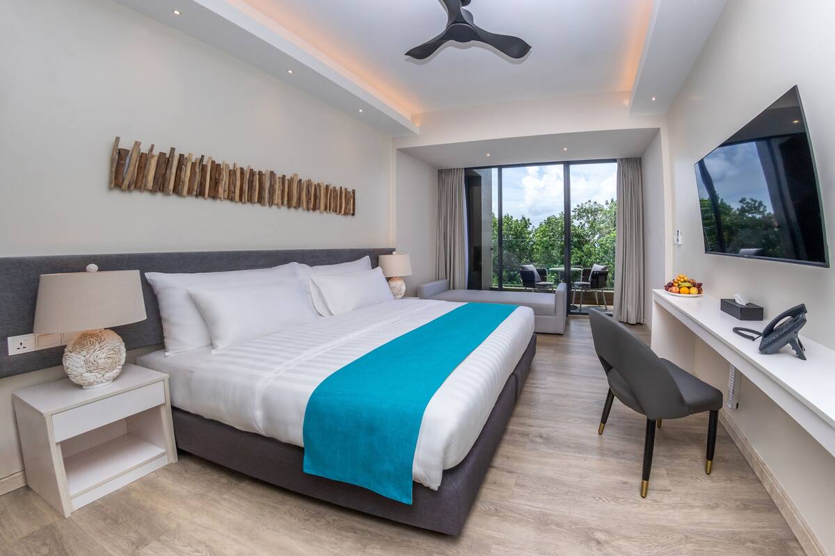 Een moderne hotelkamer met een groot bed, een bureau, een stoel, een televisie en een balkon met uitzicht op bomen.