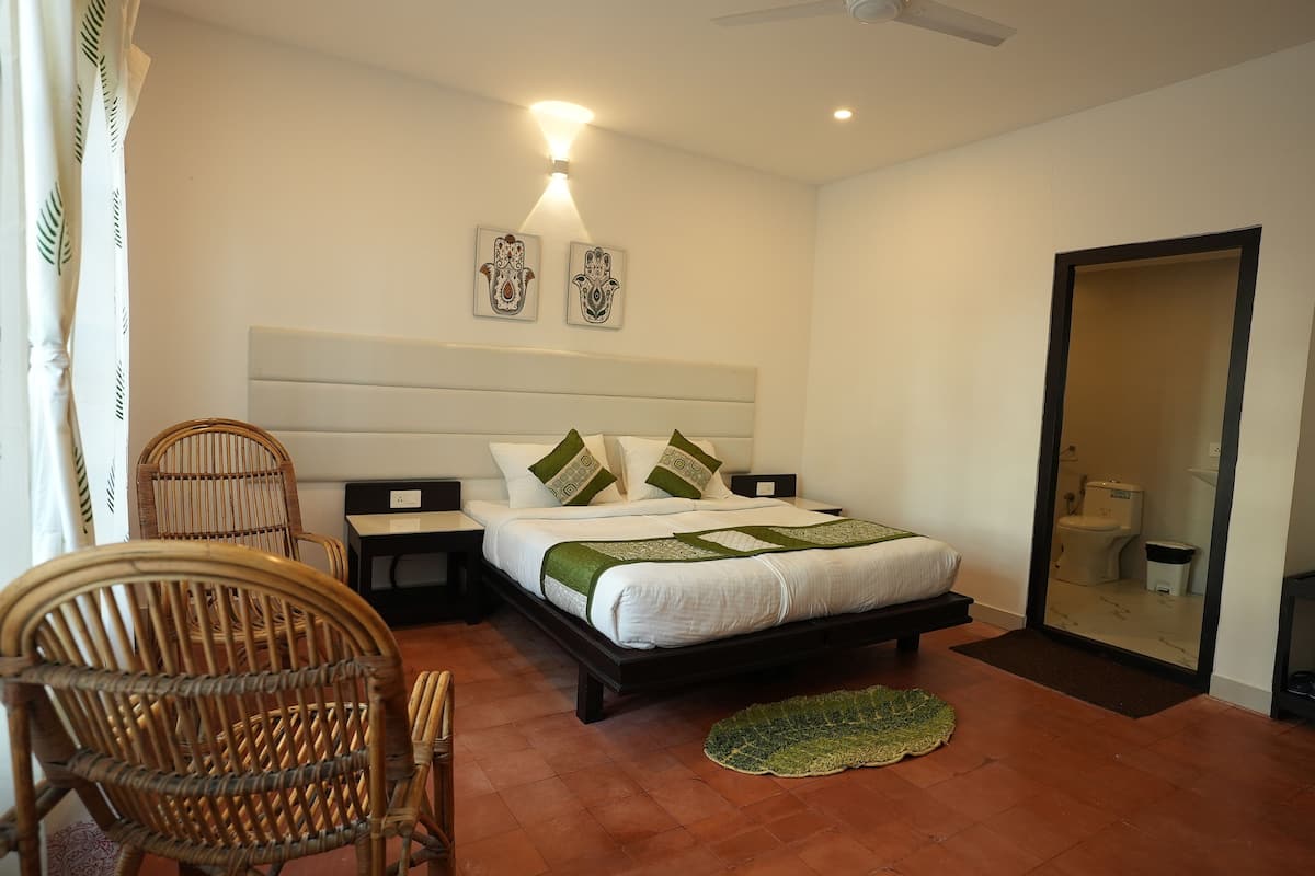 Une chambre à coucher avec un lit, deux tables de chevet, une chaise, un miroir et une salle de bain visible à travers la porte.