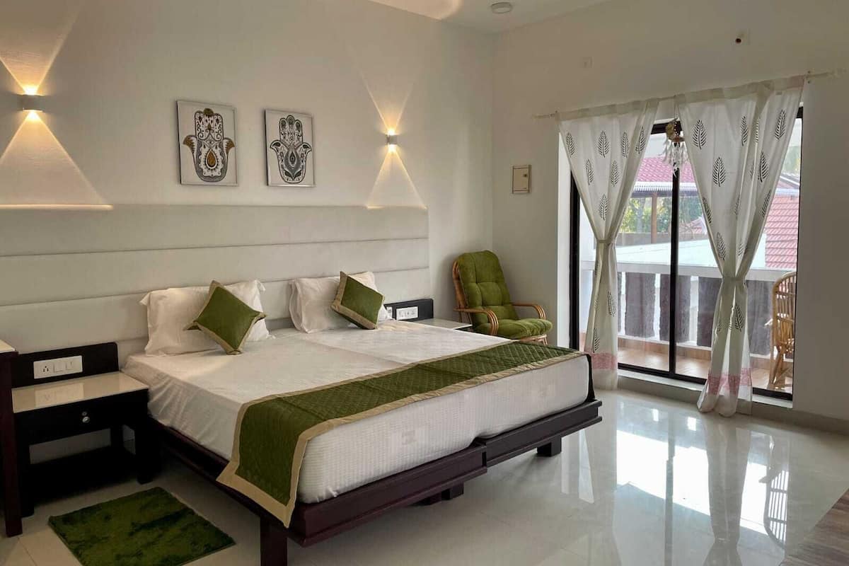 Une chambre avec un grand lit, deux tables de chevet, un fauteuil vert et une vue sur l’extérieur par une fenêtre avec des rideaux.