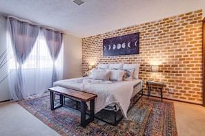 A bedroom with a brick wall, a bed with white bedding, a bench, and a colorful rug. - 🌈Tranquil condo with cozy king beds🌈decaf/ regular coffee✨work from🏡✨fast wifi✨ (Las Vegas)