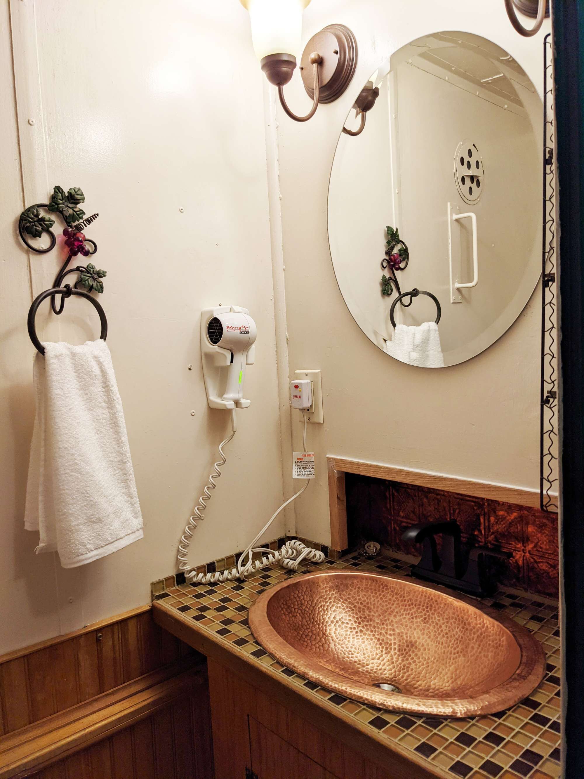 A bathroom with a copper sink, a towel rack with a white towel, a hairdryer, and a mirror with a clock.