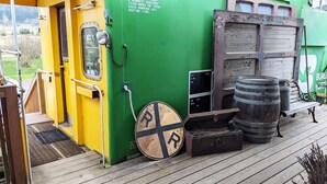 A green and yellow train car with a door, a round sign with a cross, and two barrels on a wooden deck. - Steampunk Train at Olympic Railway Inn - Pet Friendly (Sequim)
