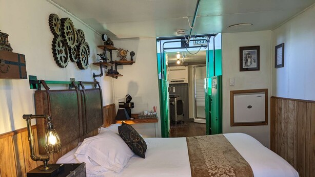 A room with a bed, a vintage lamp, a suitcase, and a green door leading to another room. - Steampunk Train at Olympic Railway Inn - Pet Friendly (Sequim)