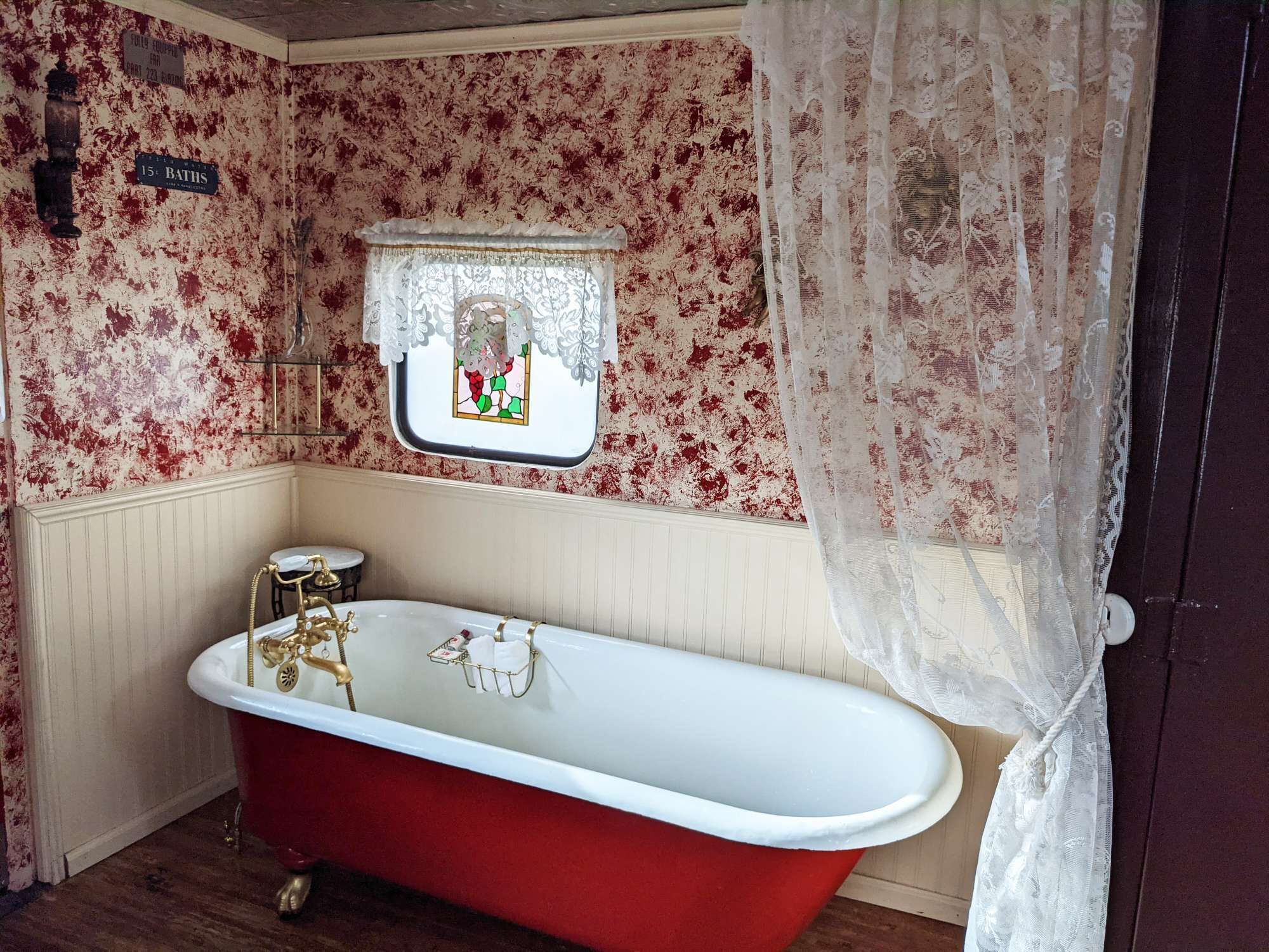 A vintage bathroom with a red clawfoot bathtub, a small window with a stained glass design, and a white lace shower curtain.