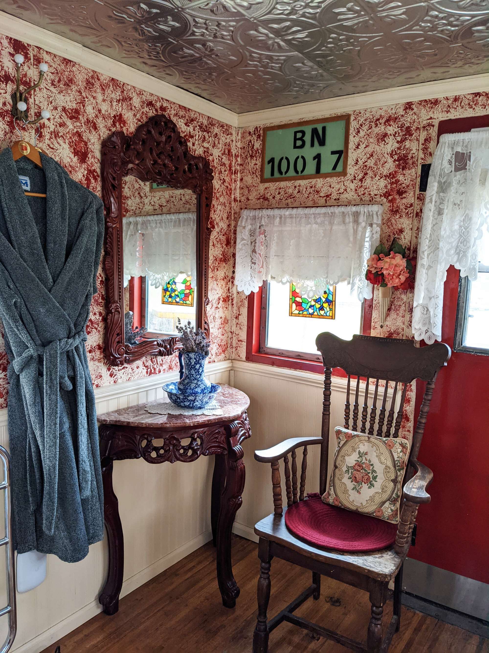 A vintage room with a wooden chair, a table with a vase, a robe hanging, and a mirror.
