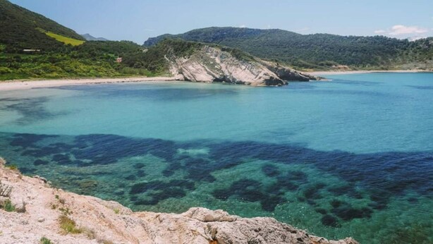Paisatge costaner amb aigües turqueses cristal·lines, roques i turons verds al fons.