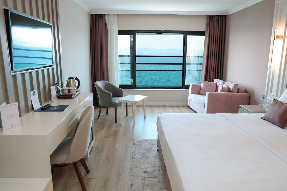 A hotel room with a bed, a desk, a chair, a sofa, and a large window with a view of the sea.