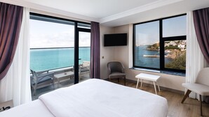 A hotel room with a large bed, a chair, a small table, and a TV. The room has large windows with a view of the sea and a coastal town. - Vira Otel Suites (Sinop)