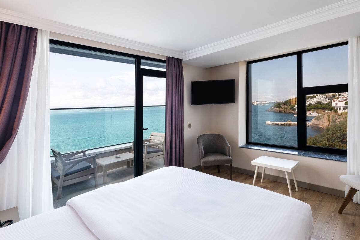 A hotel room with a large bed, a chair, a small table, and a TV. The room has large windows with a view of the sea and a coastal town.