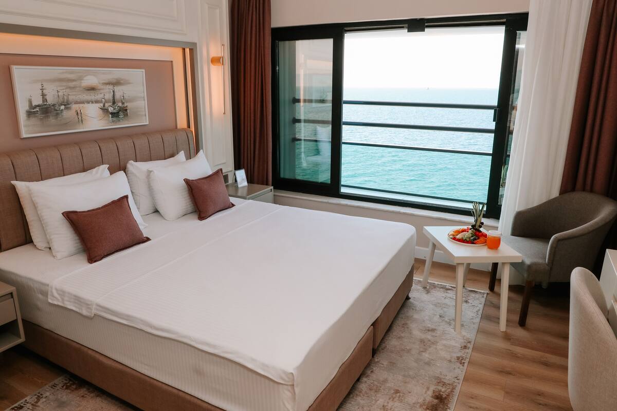 A hotel room with a large bed, a small table with a fruit bowl, a chair, and a view of the sea through a large window.