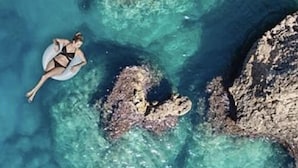 En person som flyter på en flotte i klart, blått vatten, med klippformationer i omgivningen.