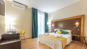 Ett hotellrum med en säng, ett nattduksbord, en lampa, en stol, ett skrivbord och en luftkonditioneringsanläggning.