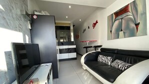 A modern living room with a black leather sofa, a flat-screen TV, and a dining area with a red table and chairs. - Charming Studio, Terrace, and Parking for Two - Steps from the Beach! (Cap d'Agde naturisme)