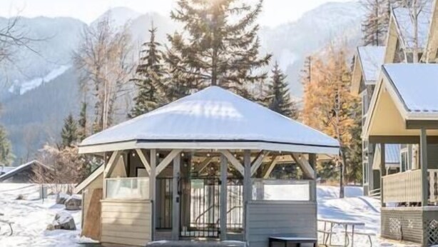 Un belvédère enneigé, doté d’une structure en bois et d’un toit en métal, entouré de neige et d’arbres, avec des montagnes en arrière-plan.