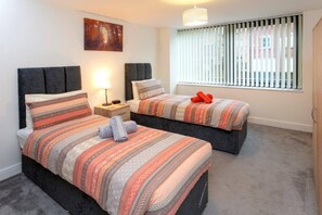 A hotel room with two beds, a nightstand, a lamp, a painting, and a window with blinds. - Station Apartment in Norwich (Norwich)