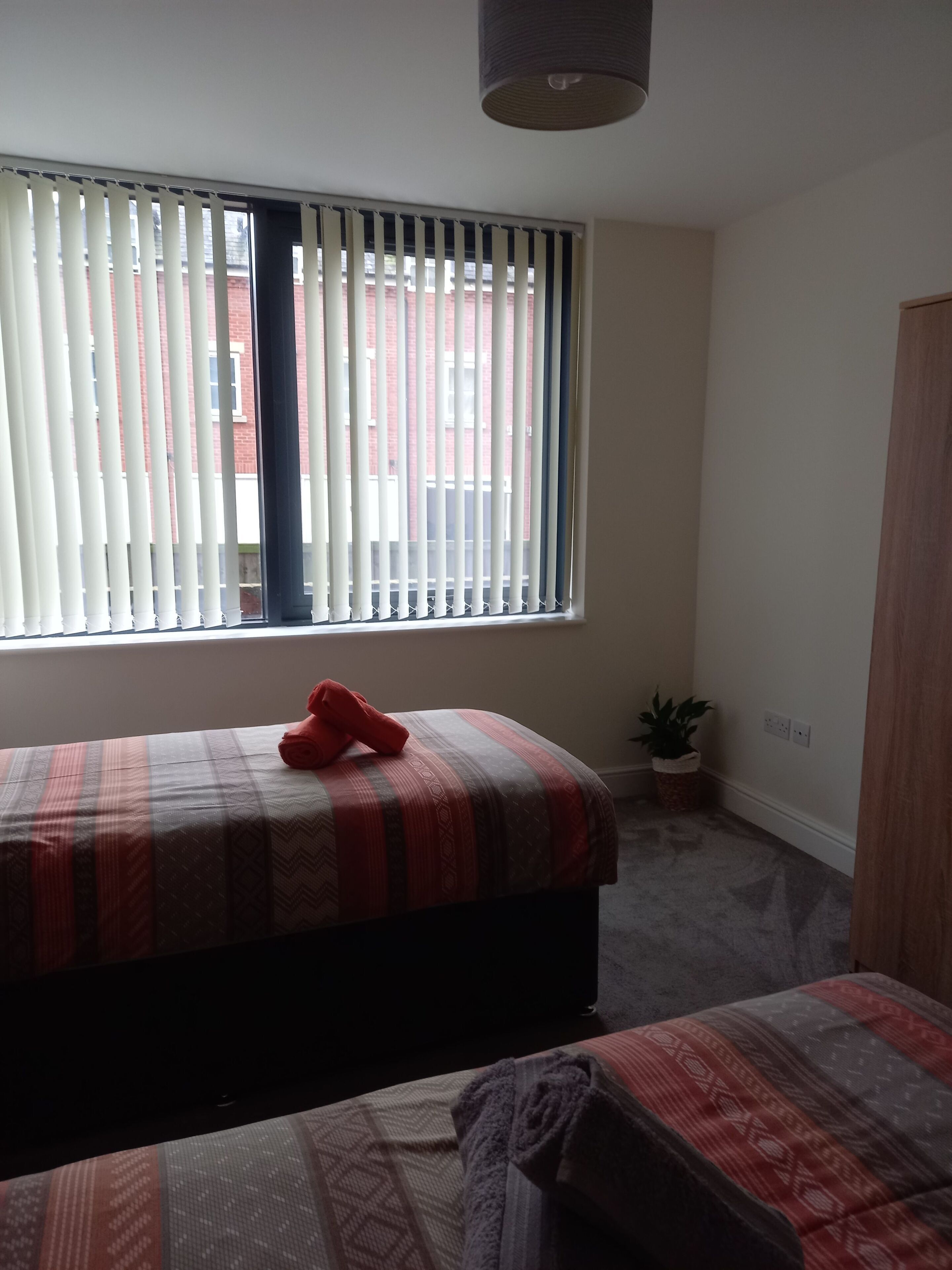 Ein Schlafzimmer mit Bett, einem Fenster mit Jalousien, einer kleinen Pflanze und einem Kleiderschrank.