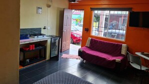 A compact kitchen with a gas stove, a refrigerator, and a sink. There is a window with a view of the street and a red car parked outside. A pink sofa is placed near the window. - Super Cheap Loft Style Apartment in Merida Wifi Smartv (Mérida)