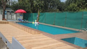 Una piscina con cubierta de madera, una sombrilla roja y un área cercada con árboles.