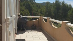Ein Balkon mit Fliesenboden, einem überdachten Grill und Blick auf einen bewaldeten Hang.