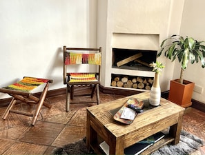 Une chaise en bois avec un siège et un dossier tissés aux couleurs vives, une petite table en bois sur laquelle se trouvent une télécommande et une carte, et une plante en pot placée à côté d’une cheminée.