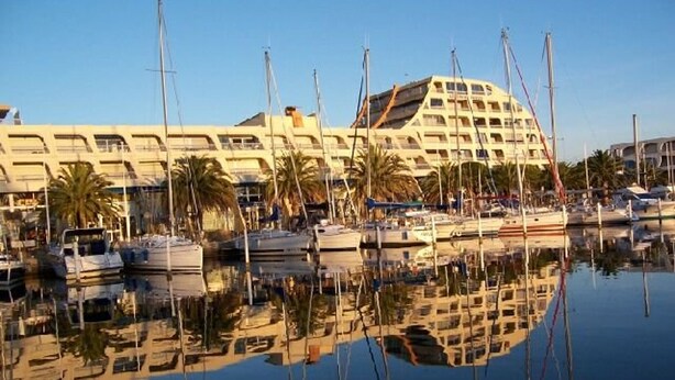 Un port de plaisance où sont amarrés de nombreux bateaux, un grand bâtiment doté de balcons et des palmiers.