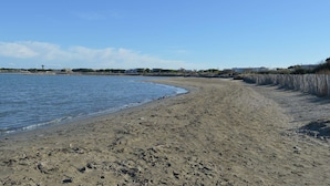 Une plage de sable bordée d’une eau d’un bleu limpide, une clôture en bois et des bâtiments visibles à l’arrière-plan.