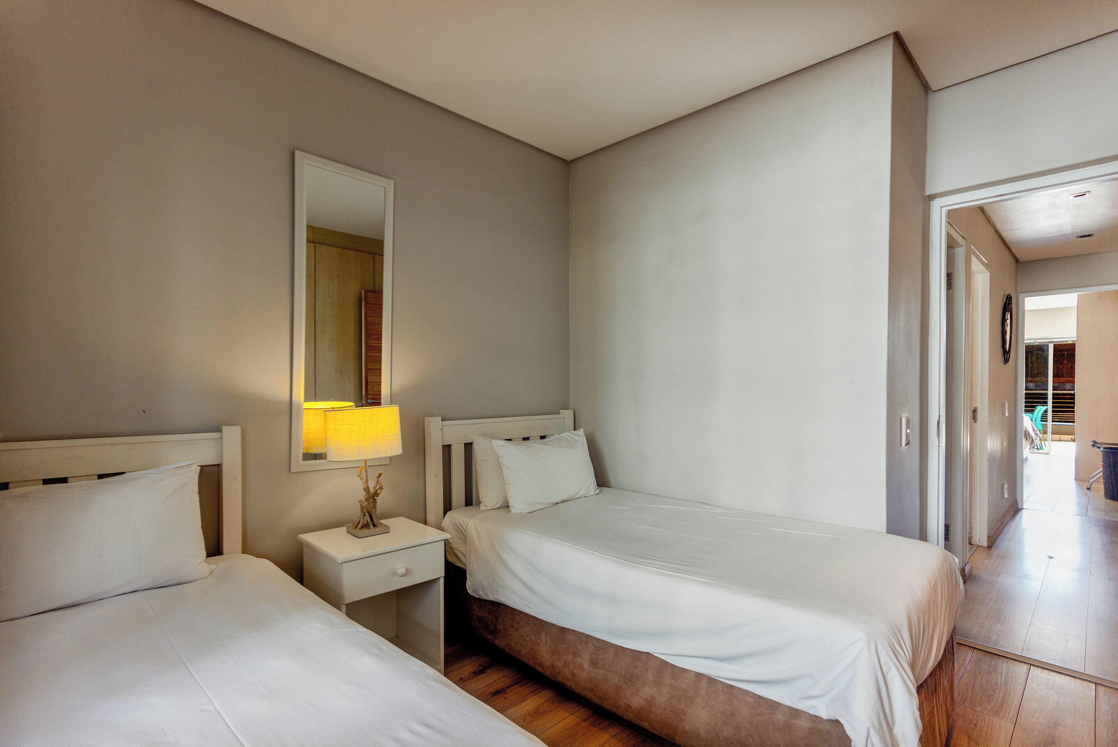 Quarto de hotel com duas camas, uma mesa de cabeceira com abajur, um espelho e chão de madeira.