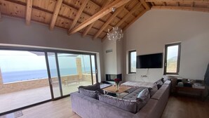 A spacious living room with a large gray sectional sofa, a wooden floor, and a large sliding glass door leading to a balcony with a view of the sea. - Luxury Villa With Private Pool Kika Residences (West Mani)