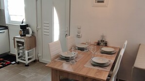 Un coin repas avec une table en bois dressée pour quatre personnes, entourée de chaises blanches. On aperçoit une machine à café et un petit meuble en arrière-plan.