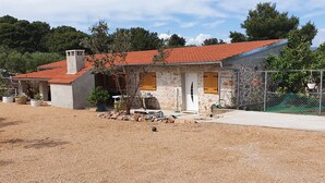 Une maison en pierre avec un toit recouvert de tuiles rouges, entourée de gravier, de plantes en pot et d’un espace délimité par une clôture, où sont suspendues des jardinières.