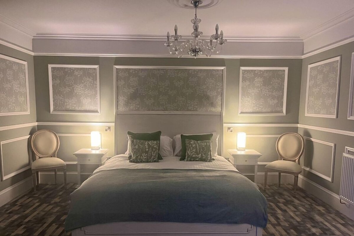 A bedroom with a large bed, two bedside tables, a chandelier, and framed floral wall art.