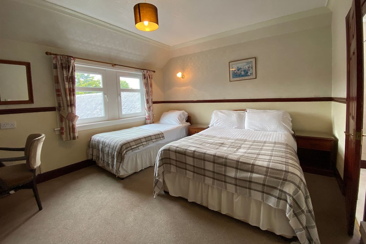 A hotel room with two beds, a desk, a chair, a window with curtains, a wall-mounted light, and a framed picture.