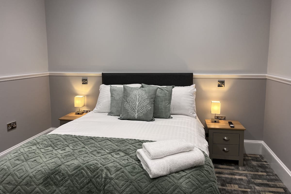 A neatly made bed with a patterned green blanket and pillows, two bedside lamps, and a nightstand with a remote control.