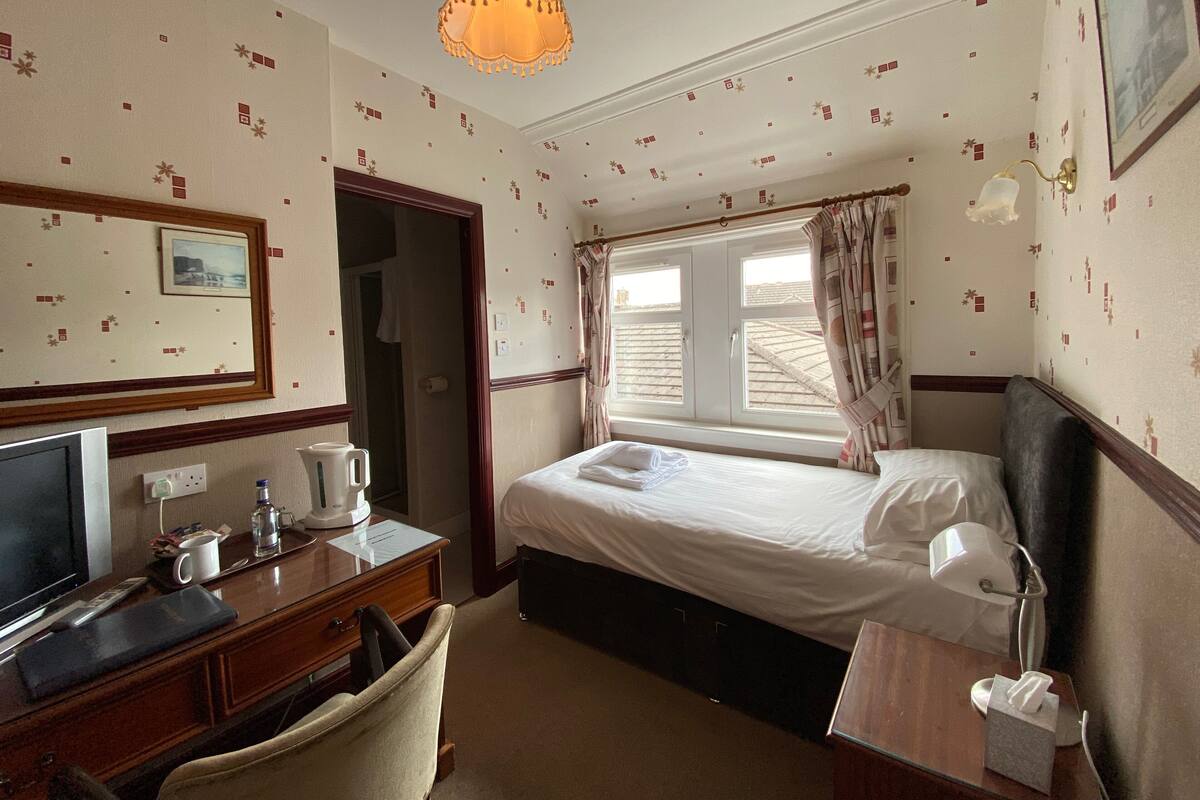 A hotel room with a bed, a desk with a television, a chair, a bedside table with a lamp, and a window with curtains.