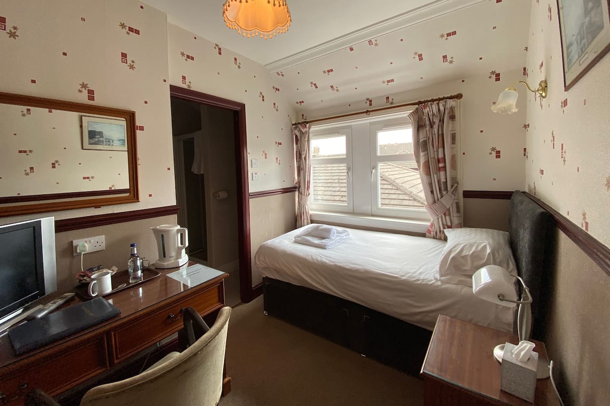 A hotel room with a bed, a desk with a television, a chair, a bedside table with a lamp, and a window with curtains.