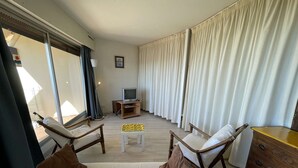 A living room with two wicker chairs, a sofa, a wooden cabinet, a television on a stand, and a window with curtains.