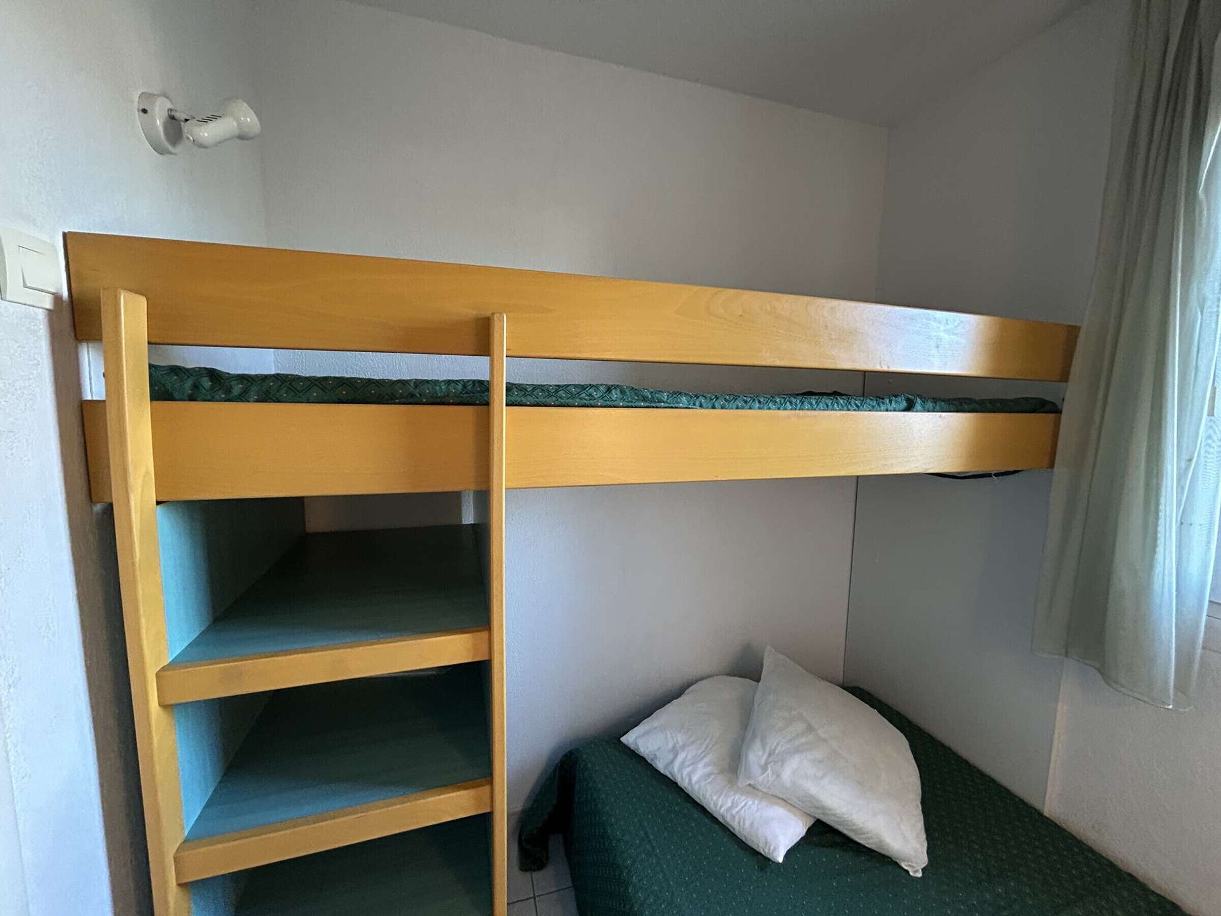 A bunk bed with a wooden frame and green bedding, a small bedside table, and a pillow.