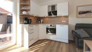 Cucina compatta con mobili bianchi, forno integrato e macchina per il caffè. Un piano di lavoro in legno e una piccola mensola con oggetti decorativi.