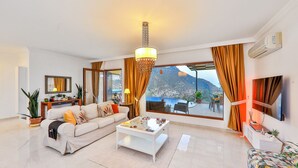 A modern living room with a sofa, coffee table, and a view of a mountainous landscape through large windows. - Villa Fiore (Antalya)
