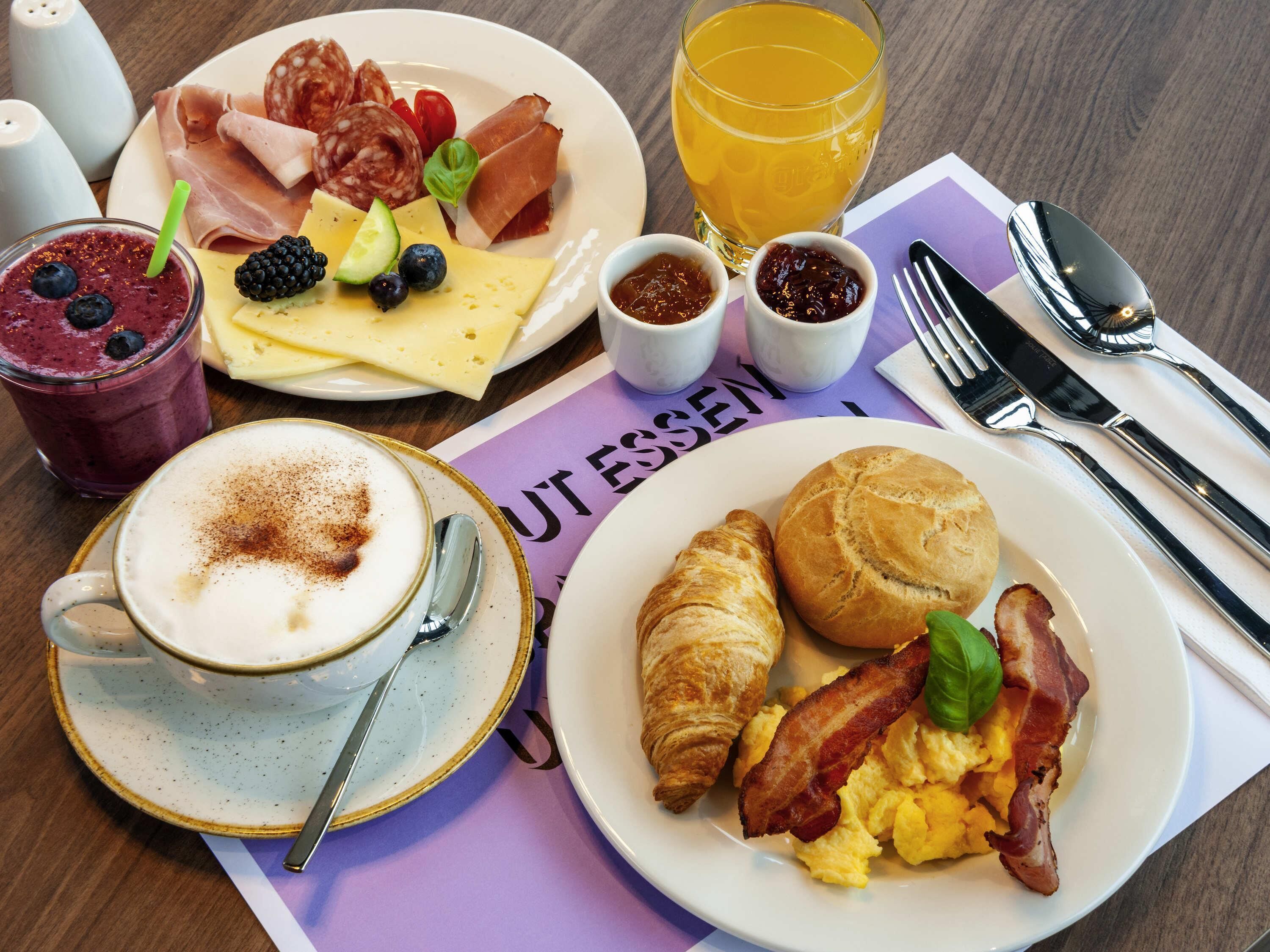 Une table de petit-déjeuner avec des croissants, du bacon, des œufs brouillés, un smoothie, du fromage et du salami.